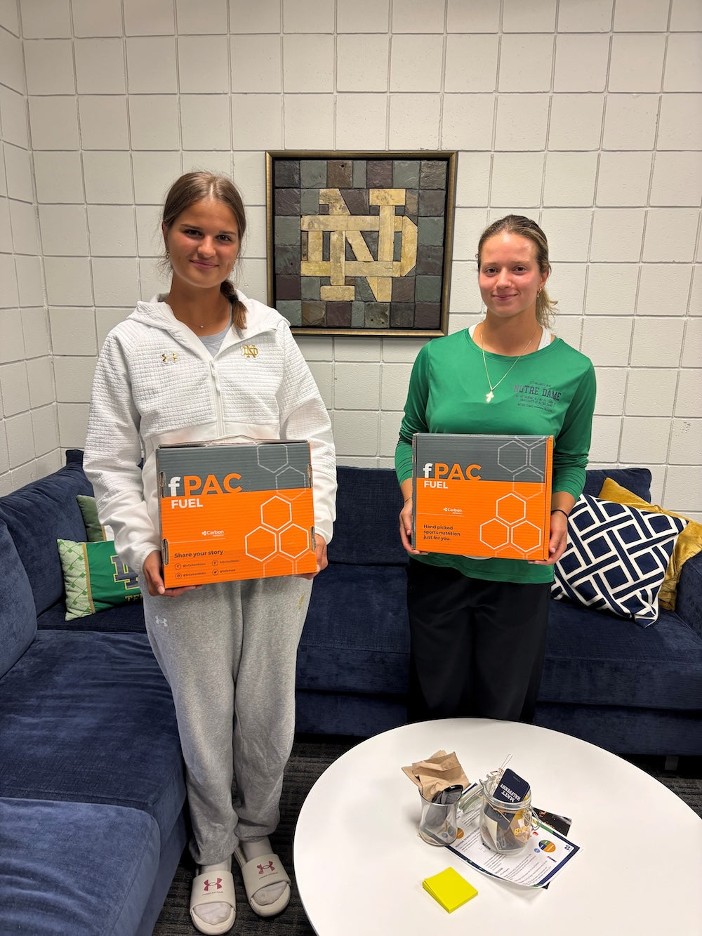 Two people holding orange boxes labeled 'FPAC' in a room with a blue couch and framed letter 'N'.