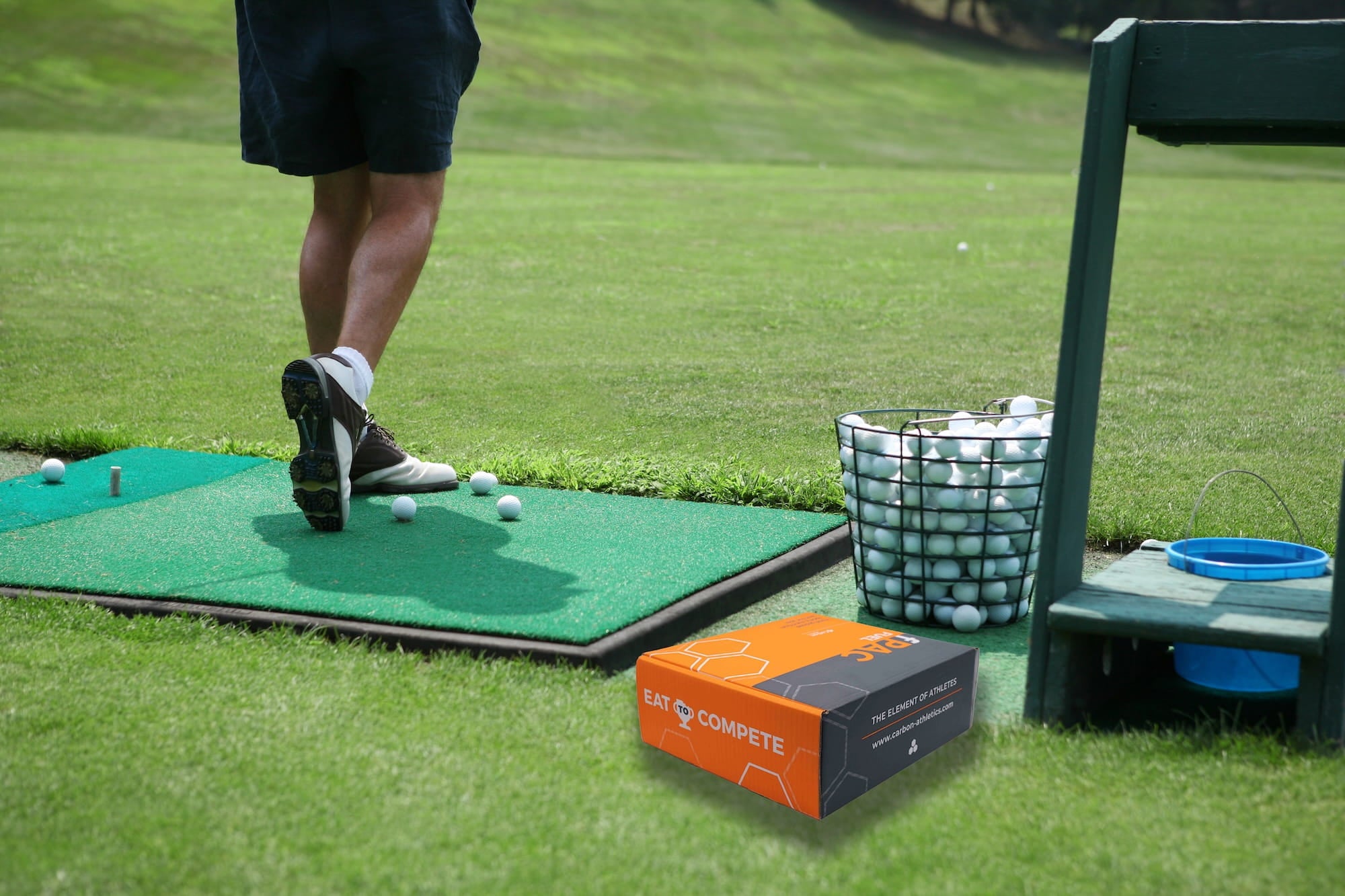 Golf practice setup with a person on a mat, golf balls, and a box on a grassy area.