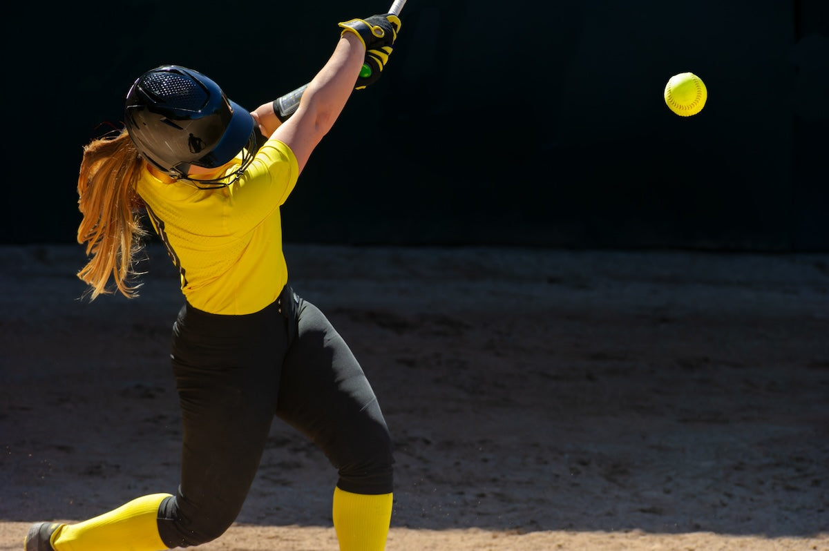 Person in yellow and black softball uniform swinging a bat at a yellow softball on a dark background