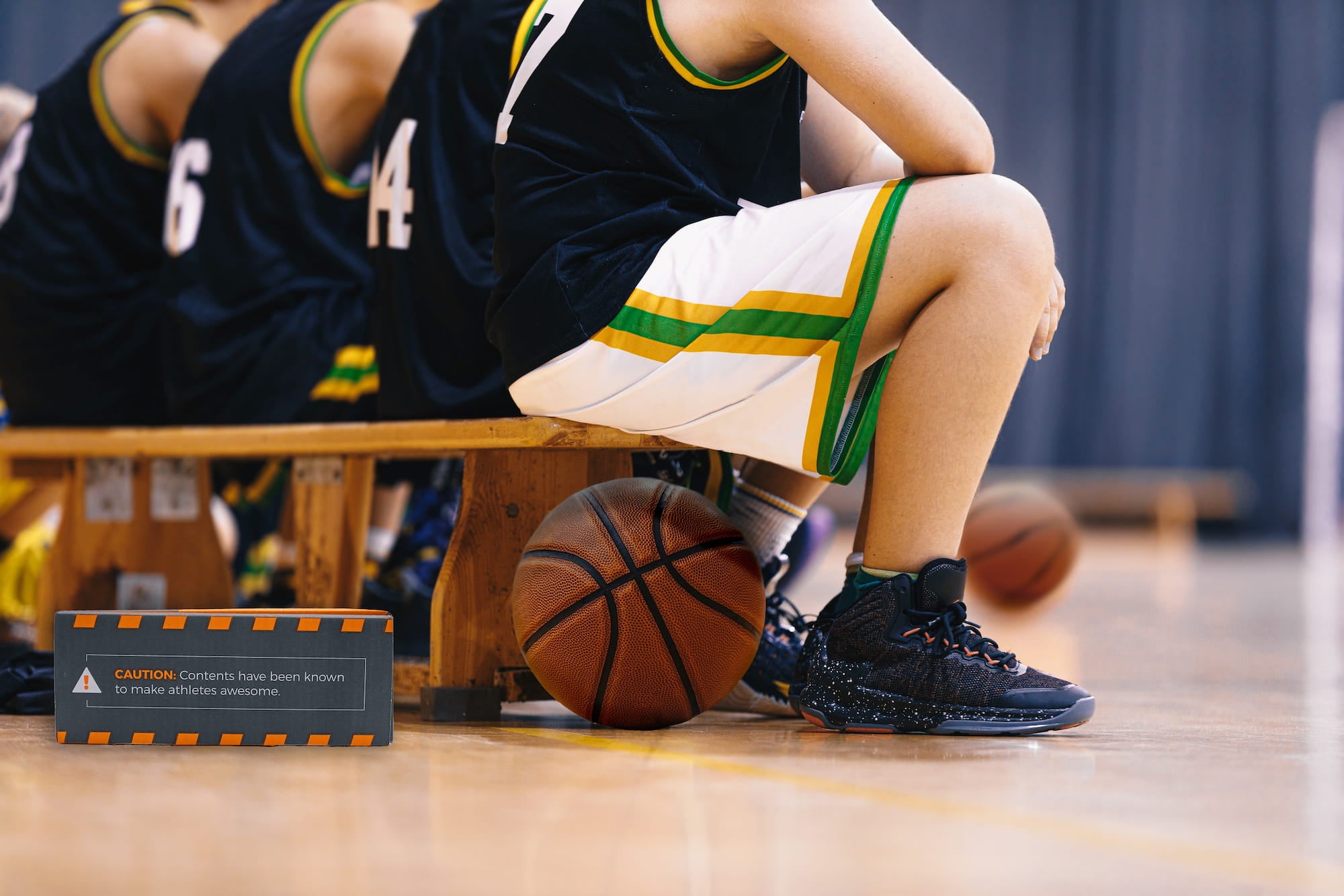 Child in basketball uniform sitting on a bench with a basketball, wearing black shoes.