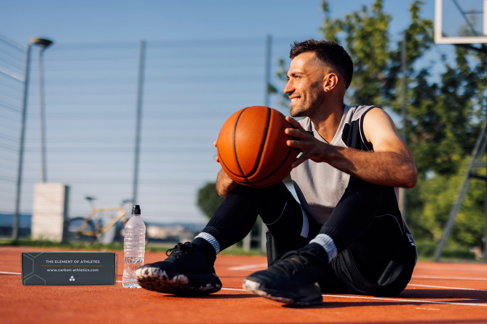 Man sitting on a basketball court holding a basketball with a water bottle beside him.
