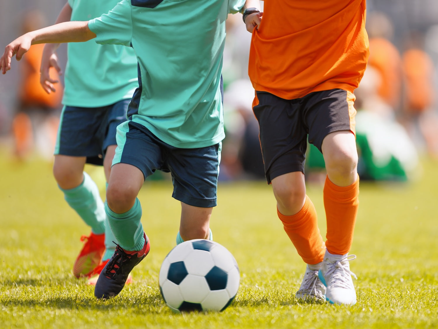 Children playing soccer on a grass field with a focus on their legs and the ball.