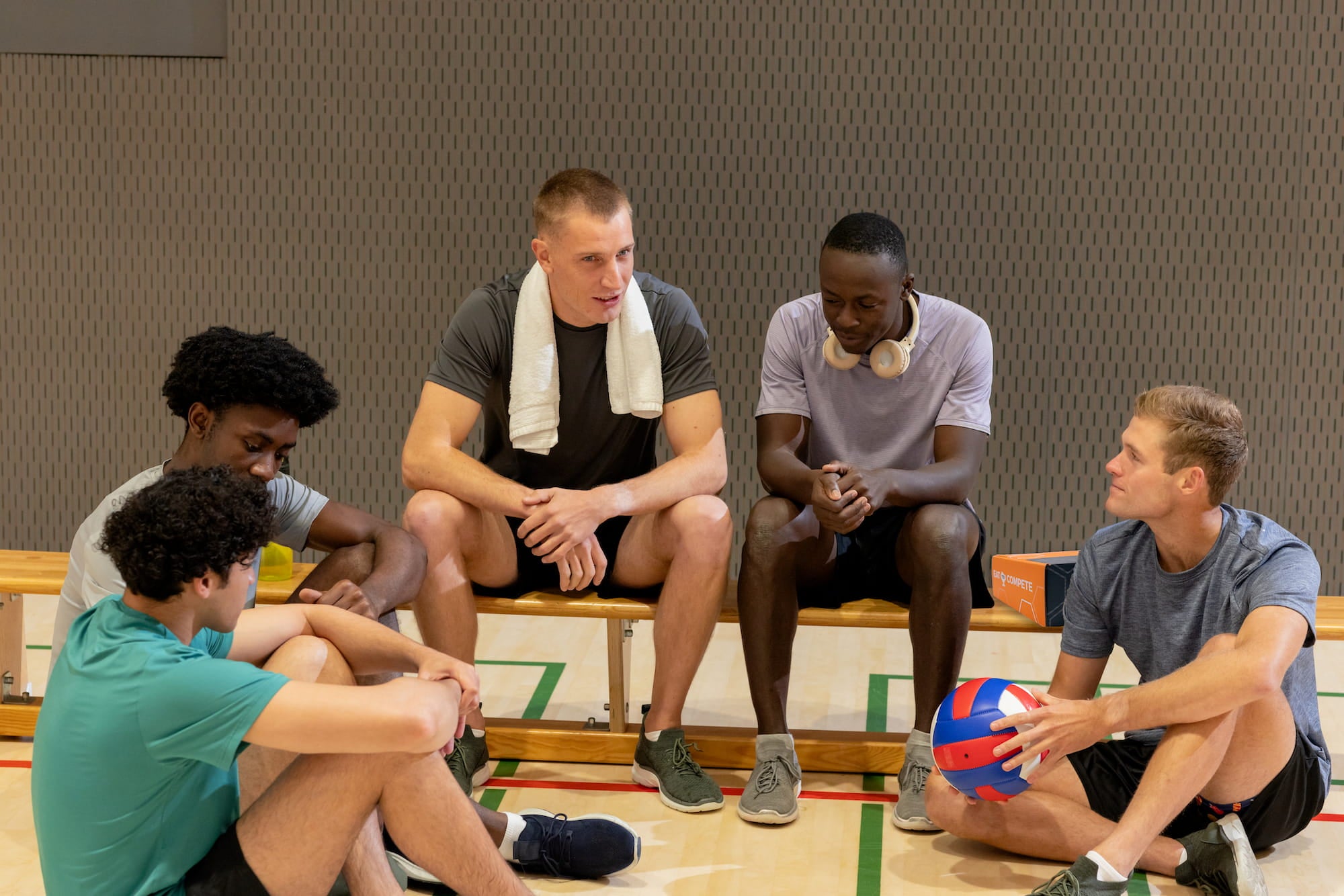 Group of young men sitting on a basketball court with sports equipment.