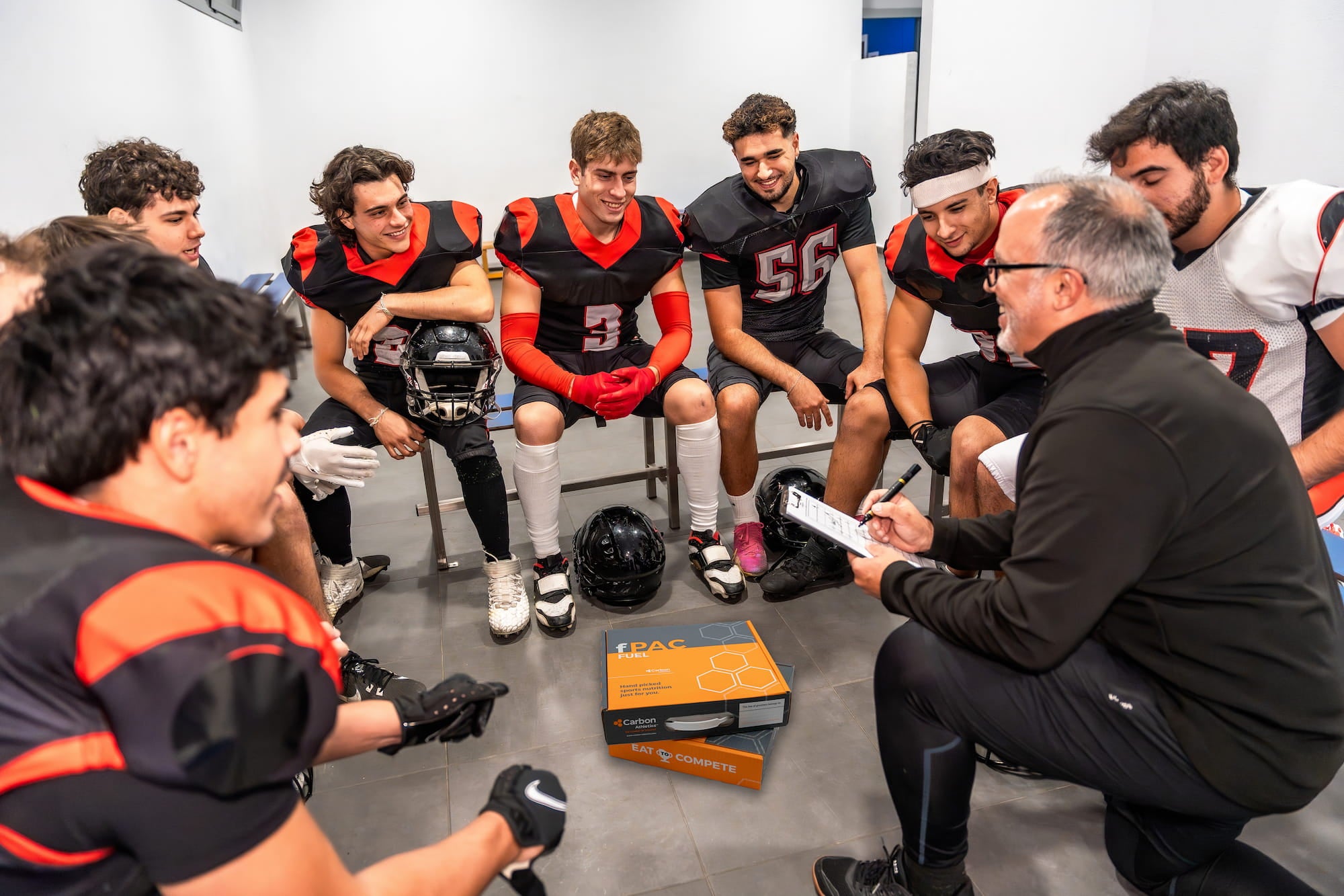 Coach with a clipboard talking to a group of football players in a locker room.