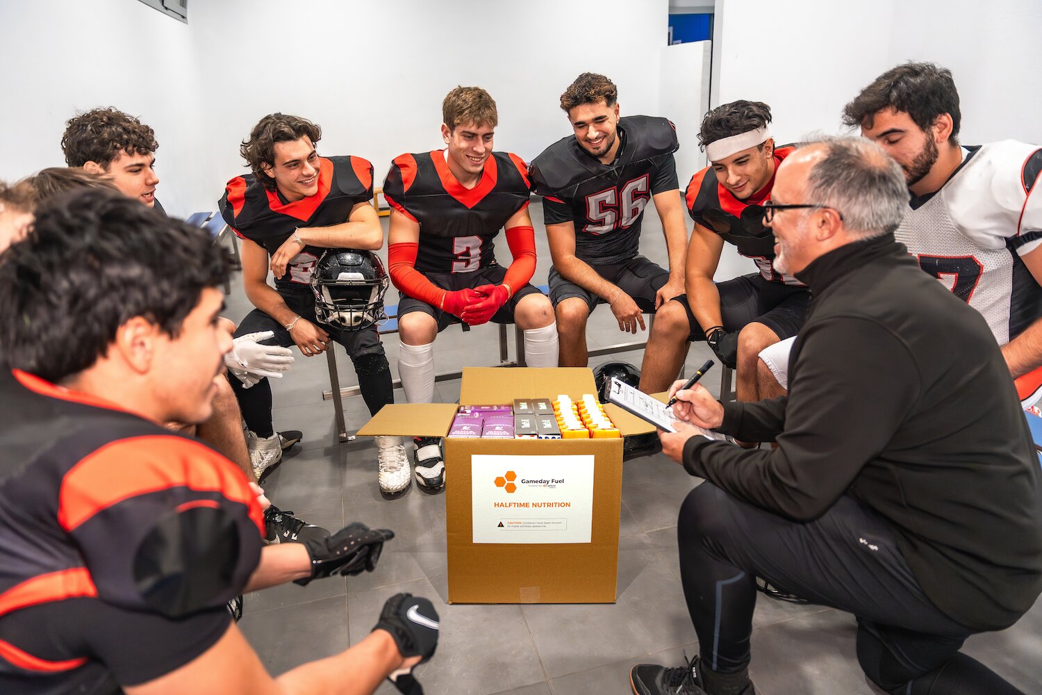 Coach with a box of sports equipment surrounded by football players in a locker room.