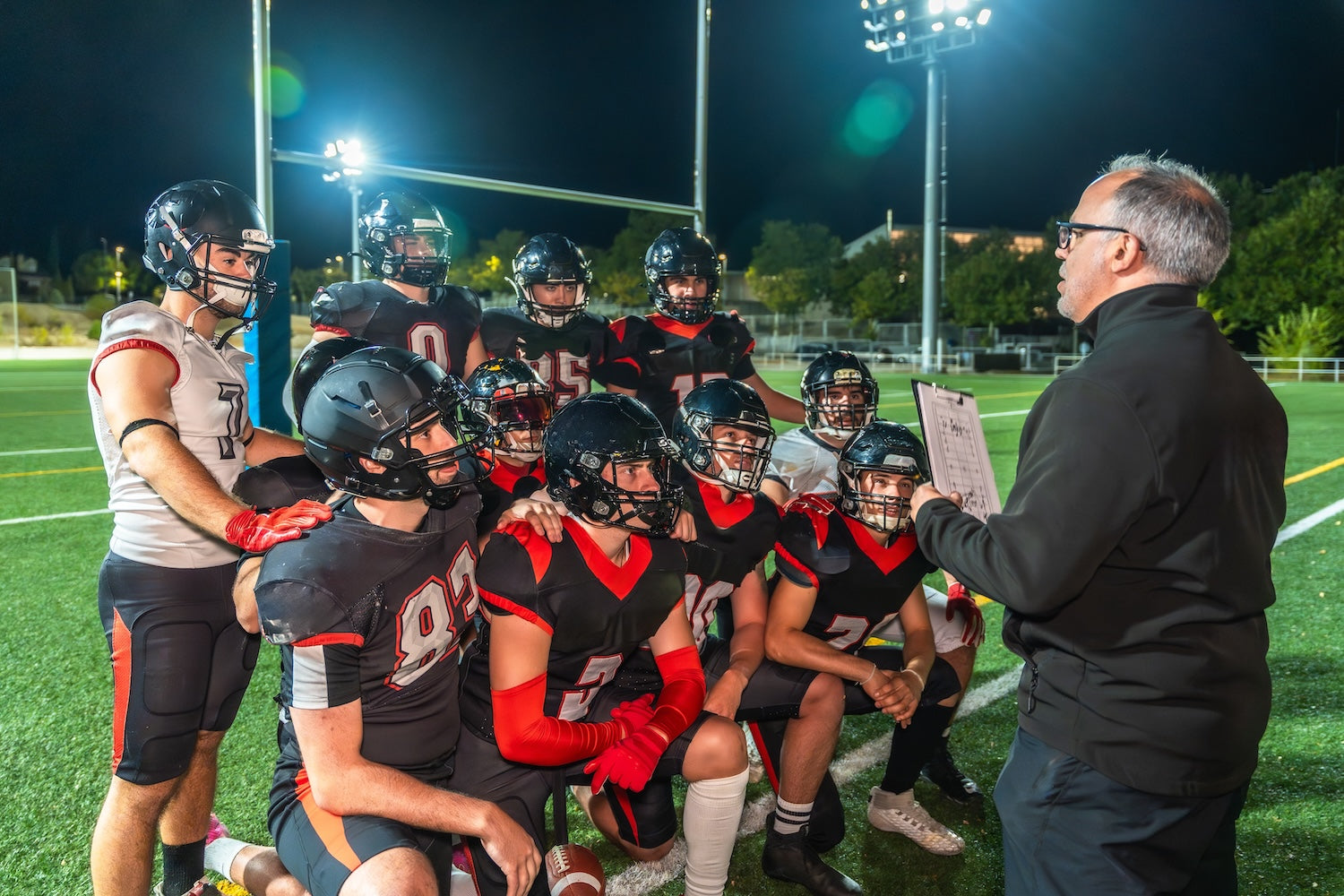 Coach giving instructions to a group of football players on the field at night.