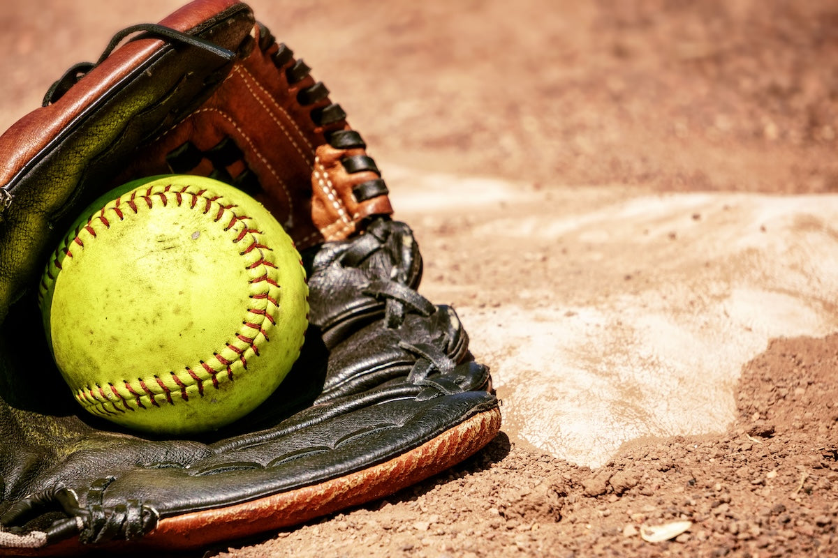 Yellow softball in a brown leather glove on a baseball field